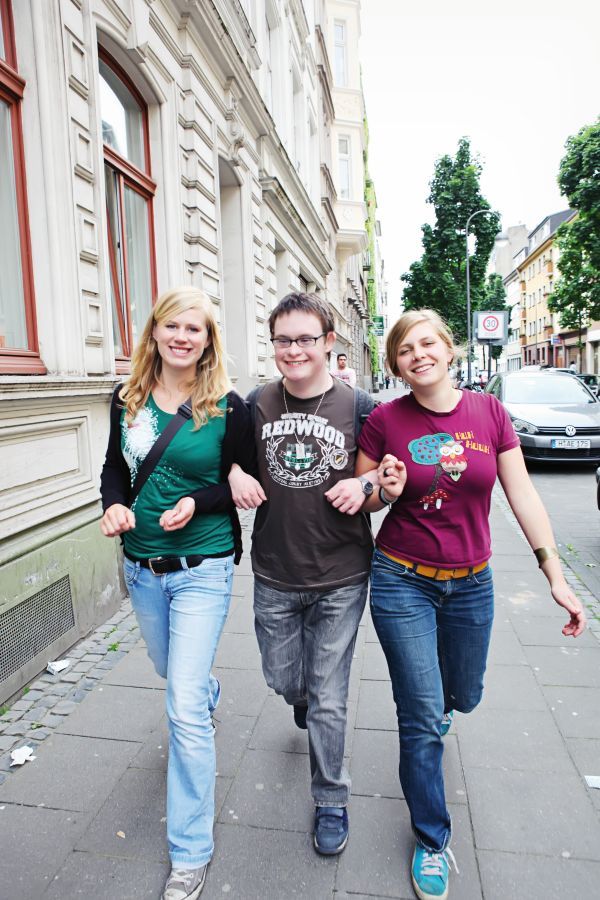 Drei junge Menschen in Jeans und bunten T-Shirts gehen Arm in Arm eine Straße hinunter.