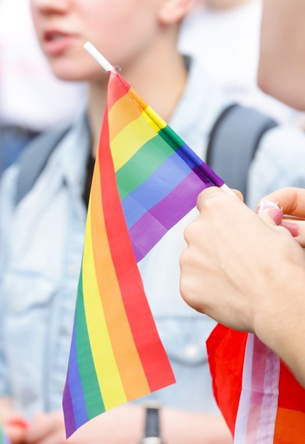 Eine junge Person hält eine kleine Regenbogenfahne in der Hand. Im Hintergrund sind Menschen zu sehen, die sich unterhalten.