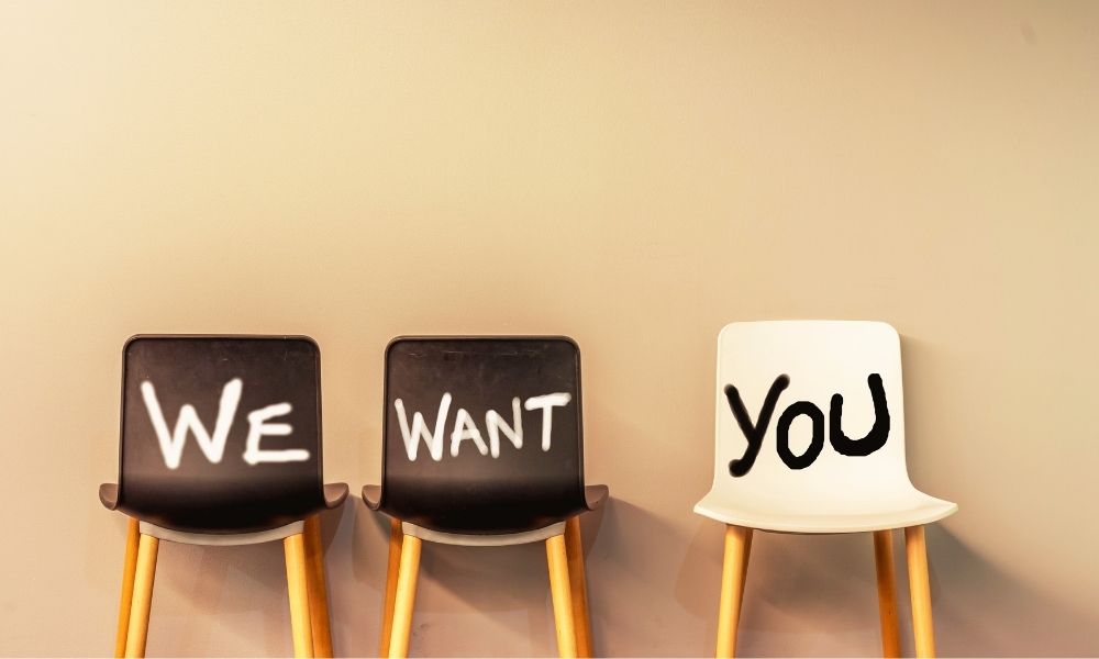 vor einer weißen Wand stehen zwei schwarze und ein weißer Stuhl, auf denen jeweils ein Wort steht: we want you