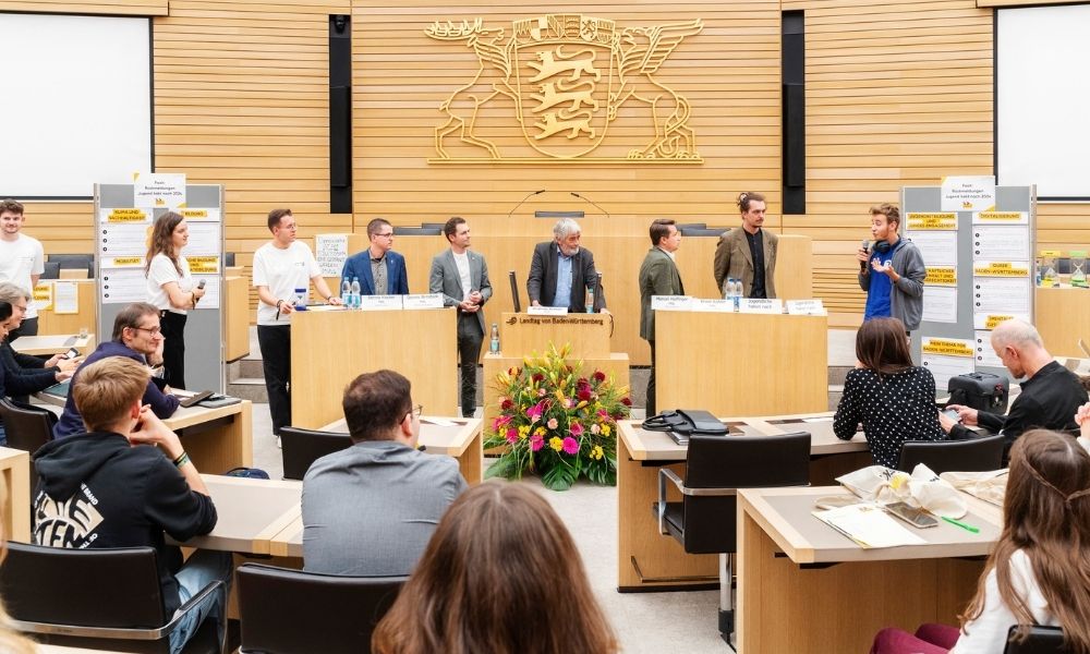 Viele Jugendliche sitzen im Plenarsaal des Landtags. Ein junger Mann moderiert am Sprecherpult, neben ihm stehen die vier Jugendpolitischen Sprecher der Legislatur 2021-2026.