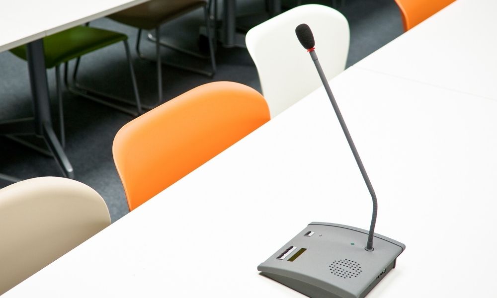 auf einem weißen Tisch steht ein Tischmikrofon, im Hintergrund ist ein orangefarbener Stuhl zwischen vielen weißen Stühlen.