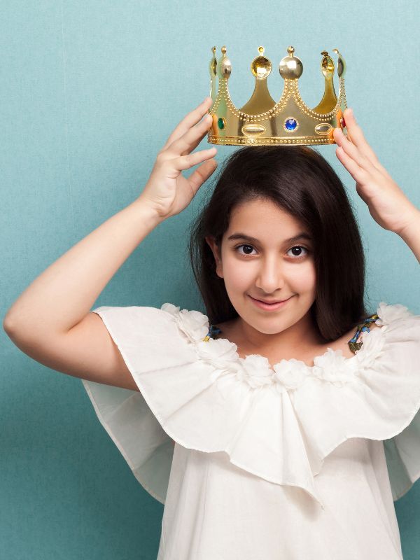 Ein Mädchen mit dunklen Haaren setzt sich lächelnd eine Krone auf.