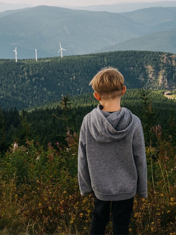 Ein Junge steht auf einem Berg, mit der Aussicht auf eine grüne, bergige Landschaft. Im Hintergrund sind mehrere Windräder zu sehen.