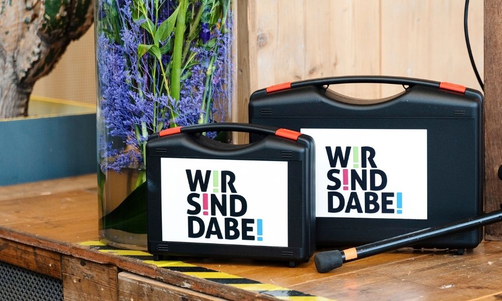 Auf einer Bühne stehen ein großer und ein kleiner Werkzeugkoffer, auf denen jeweils das Projektlogo von Wir sind dabei aufgeklebt ist.