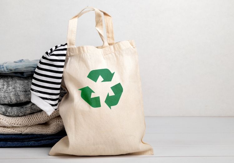 Ein kleiner Stapel Kleidung im Hintergrund, davor steht ein Stoffbeutel, auf dem das Recycling-Symbol aufgedruckt ist.